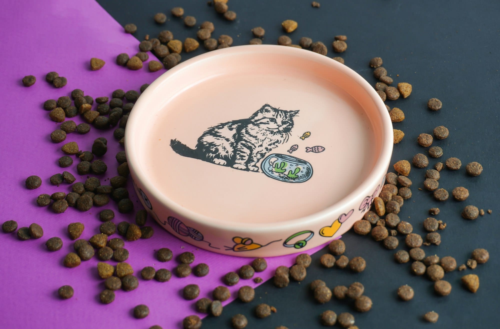 An empty pink ceramic cat bowl decorated with a kitten drawing, surrounded by scattered dry kibble